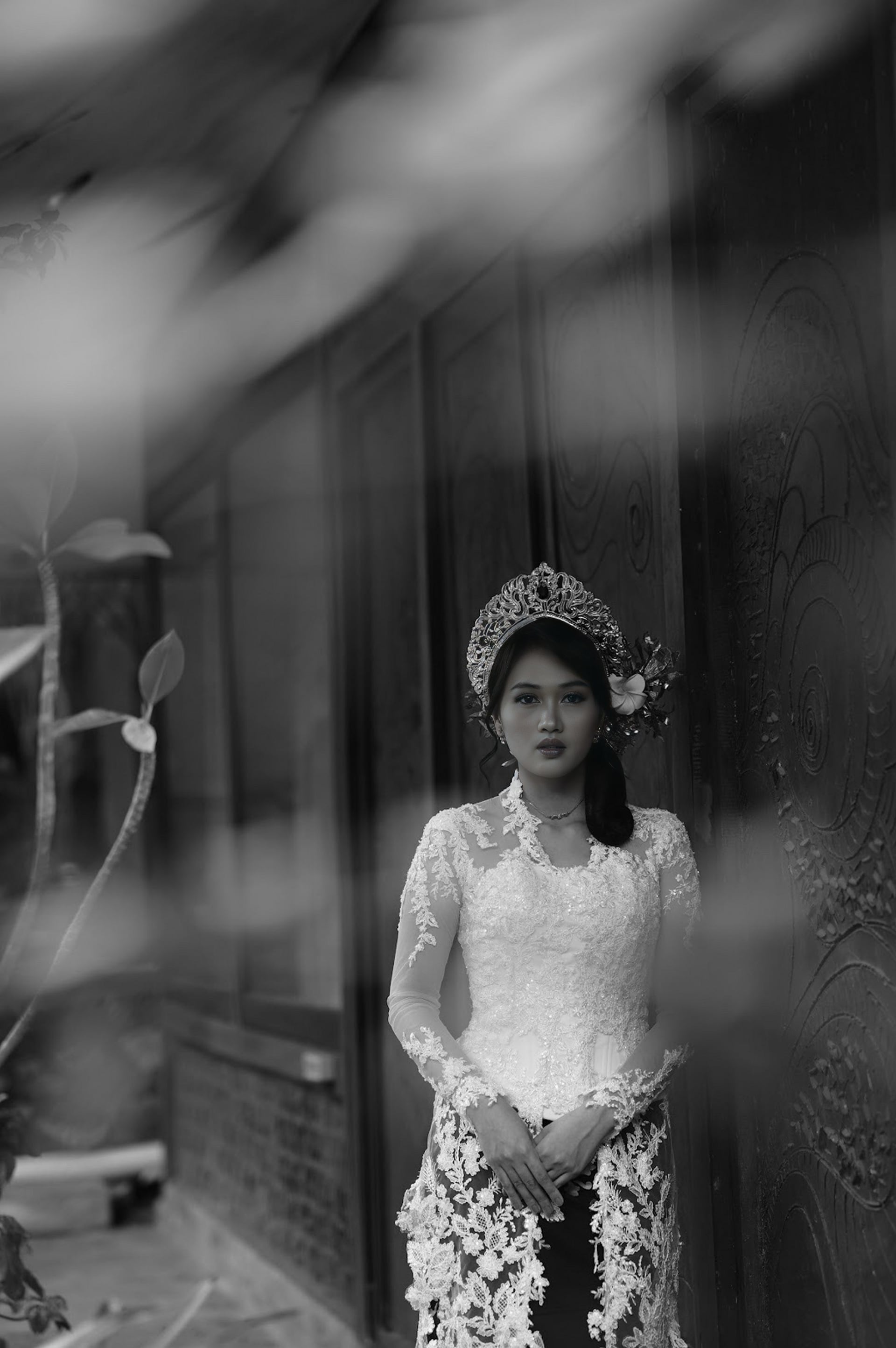 A bride in a beaded kebaya and gold crown, photographed through leaves.