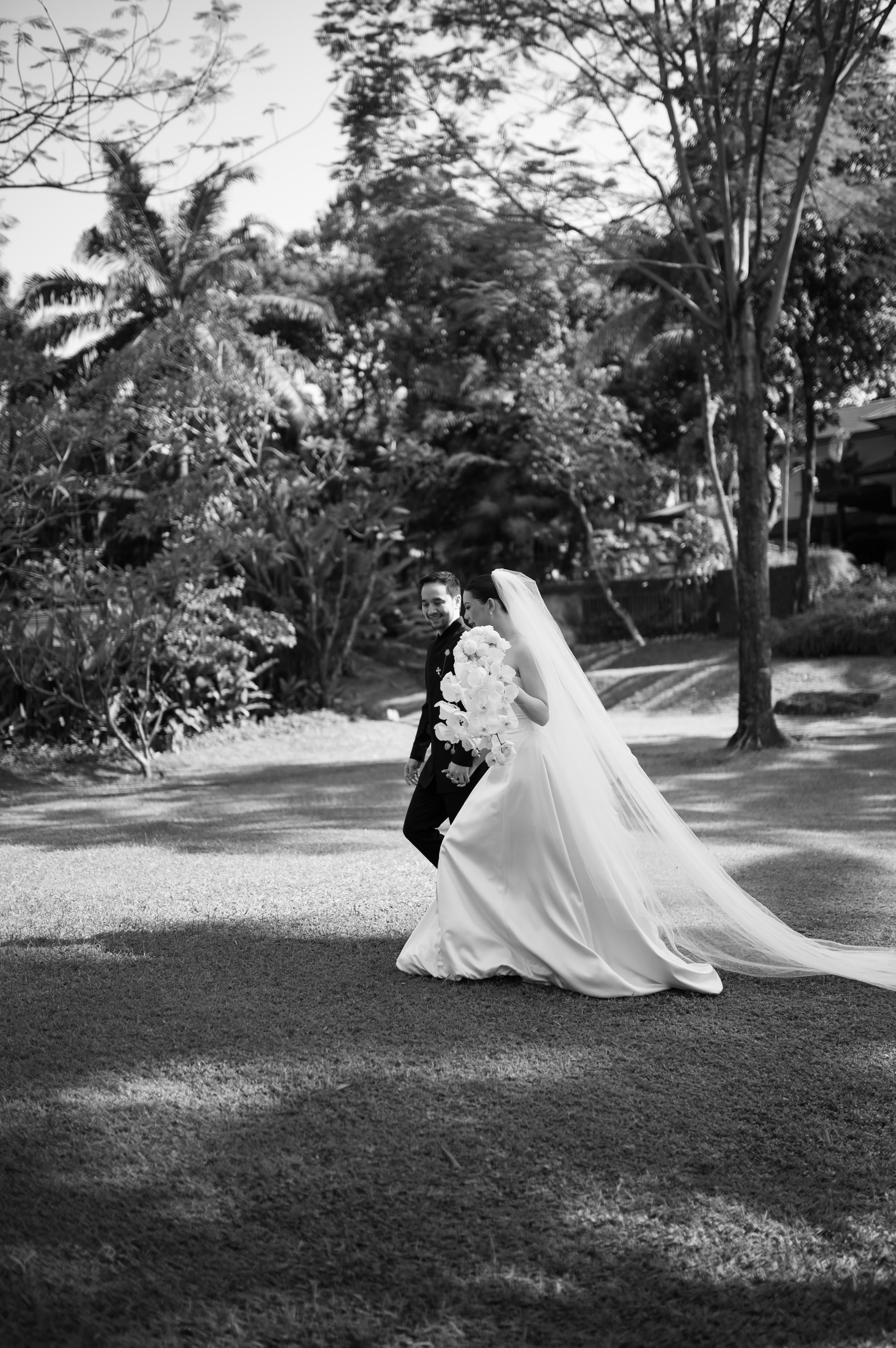 A bride walking beneath trees in a sculptural gown and long veil.