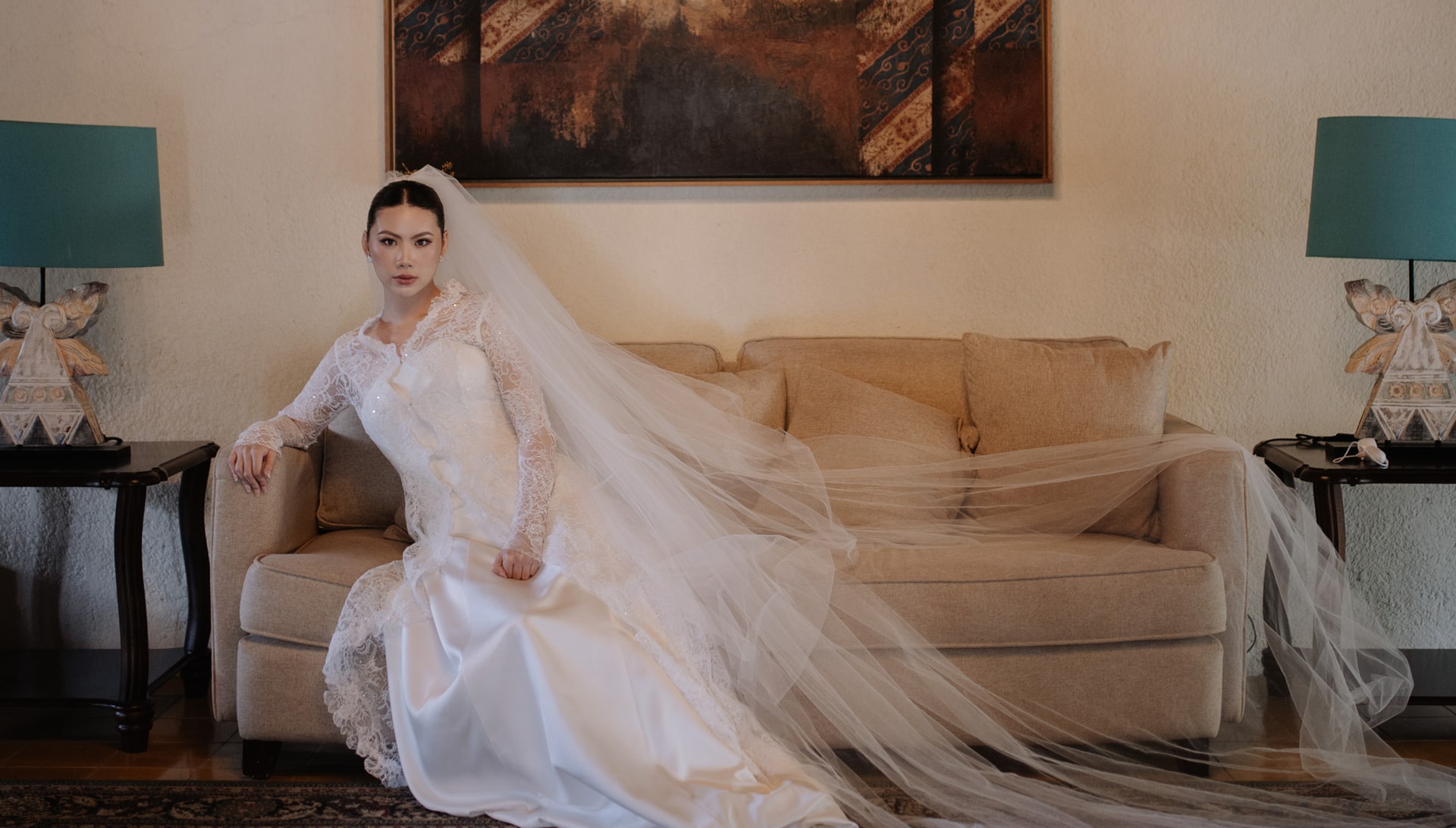 A bride in a lace gown seated beneath a large artwork before the ceremony.